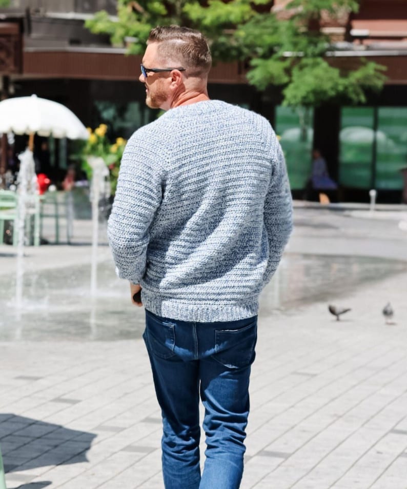 May include: A light blue sweater with a ribbed knit design. The sweater is paired with dark blue jeans. The person is wearing sunglasses and walking outdoors on a sunny day.
