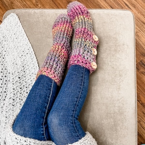 May include: A pair of colourful crocheted slipper socks with wooden buttons. The socks are pink, purple, and grey with a multi-coloured yarn. The socks are worn by a person sitting on a light grey sofa.