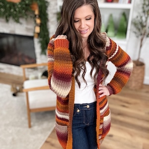 May include: A long, open-front cardigan in a warm color palette of orange, brown, and cream. The knitted sweater features horizontal stripes and a hooded design. The sleeves are long, and the cardigan appears to be made of a soft, textured yarn.