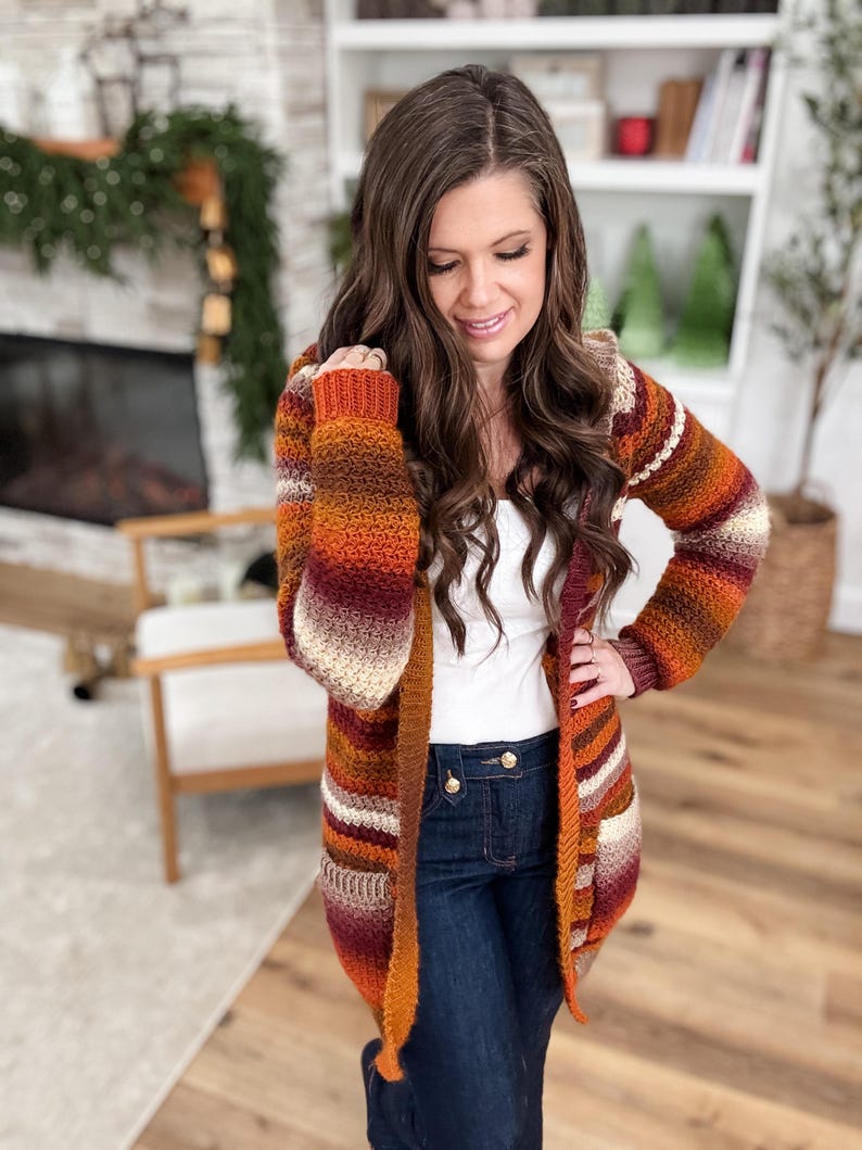 May include: A long, open-front cardigan in a warm color palette of orange, brown, and cream. The knitted sweater features horizontal stripes and a hooded design. The sleeves are long, and the cardigan appears to be made of a soft, textured yarn.