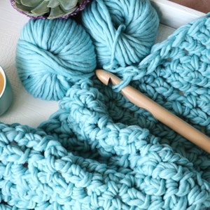 Pu&ograve; includere: Primo piano di una coperta all'uncinetto blu con un uncinetto di legno e due gomitoli di lana blu. La coperta &egrave; realizzata con un filato spesso e ha un semplice motivo a punto.