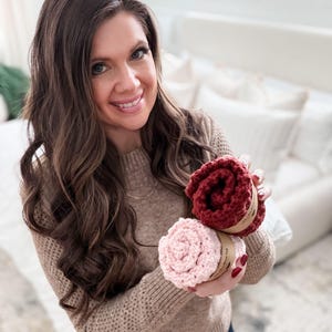 May include: A woman holding two rolled-up, circular, knitted items. One is a deep red color, and the other is a light pink. Both are wrapped with a brown paper band. The woman is wearing a tan sweater.