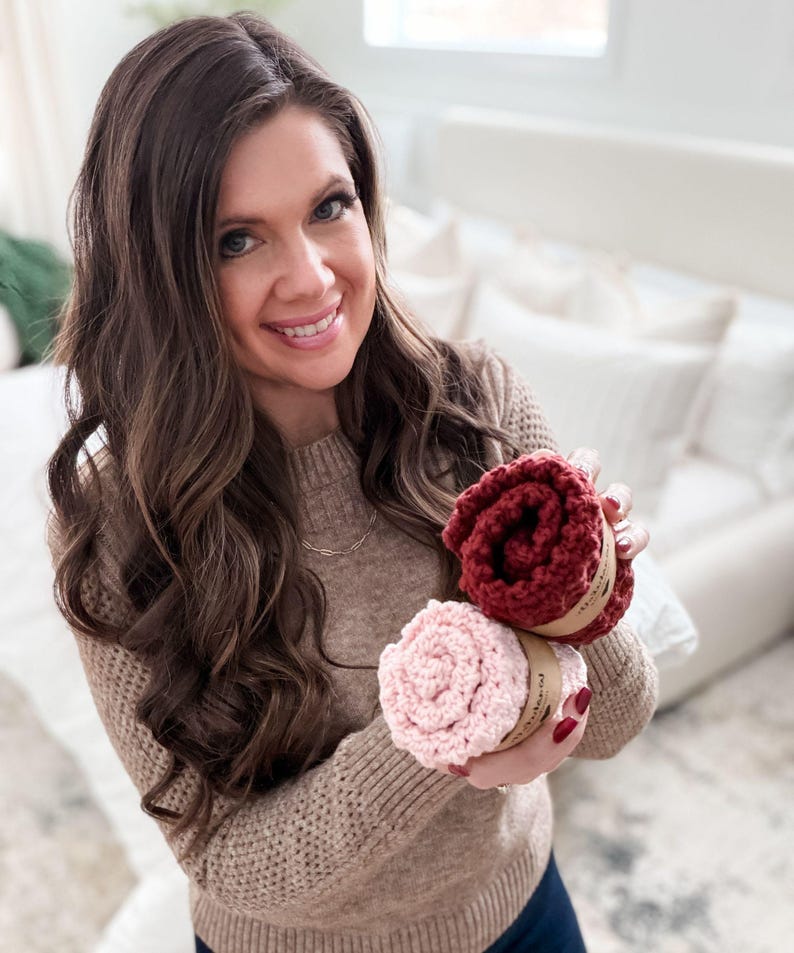 May include: A woman holding two rolled-up, circular, knitted items. One is a deep red color, and the other is a light pink. Both are wrapped with a brown paper band. The woman is wearing a tan sweater.