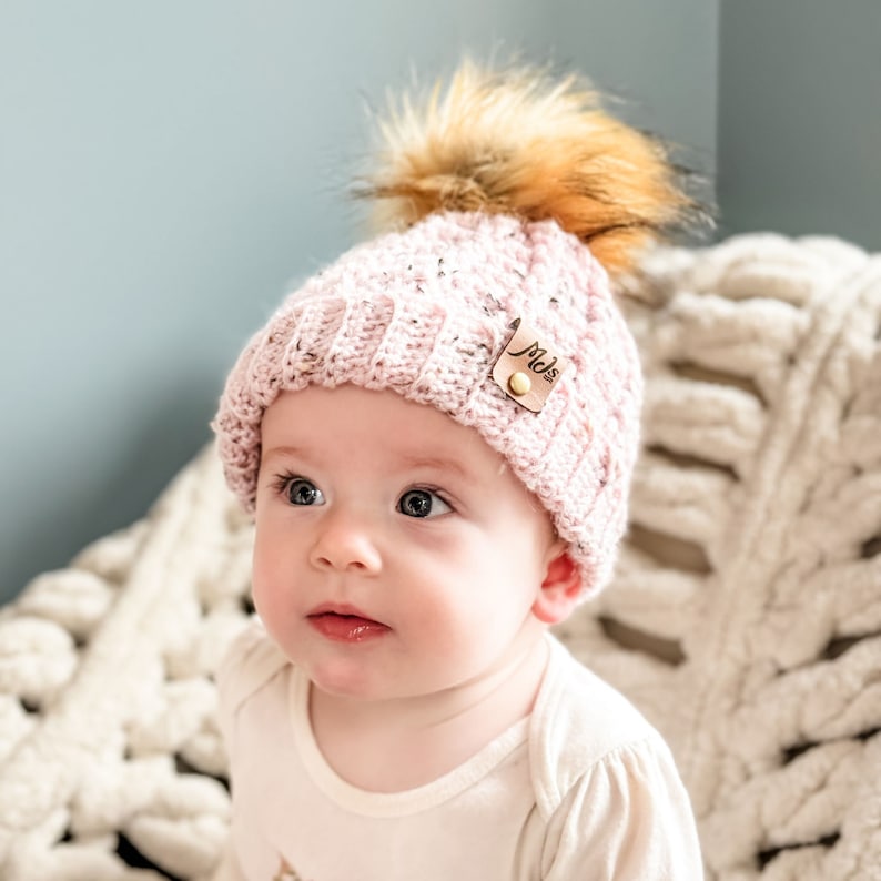 May include: A pink crocheted beanie with a faux fur pom pom and a brown leather tag that says "Mels". The beanie is on a baby's head.
