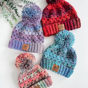 May include: Four colorful crocheted hats with pom-poms. Each hat has a brown leather tag with the text "Mels Handmade Designs".