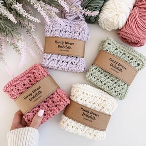 Puede incluir: Una colecci&oacute;n de pa&ntilde;os de cocina Honey Weave hechos a mano en varios colores pastel: rosa, lavanda, verde y crema. Cada pa&ntilde;o est&aacute; envuelto con una banda de papel marr&oacute;n que dice "Honey Weave Dishcloth Handmade". Madejas de hilo en colores coordinados se colocan en el fondo.