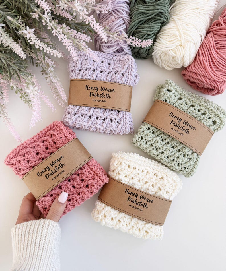 Puede incluir: Una colecci&oacute;n de pa&ntilde;os de cocina Honey Weave hechos a mano en varios colores pastel: rosa, lavanda, verde y crema. Cada pa&ntilde;o est&aacute; envuelto con una banda de papel marr&oacute;n que dice "Honey Weave Dishcloth Handmade". Madejas de hilo en colores coordinados se colocan en el fondo.