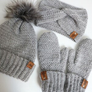 May include: A gray crocheted beanie with a faux fur pom pom, a gray crocheted headband, and a pair of gray crocheted mittens. Each item has a brown leather tag with a logo.
