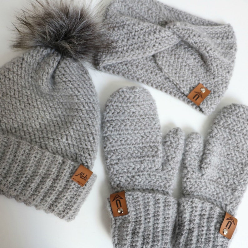 May include: A gray crocheted beanie with a faux fur pom pom, a gray crocheted headband, and a pair of gray crocheted mittens. Each item has a brown leather tag with a logo.