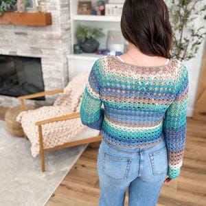 May include: A crocheted top in shades of blue, teal, and brown. The long-sleeved top has a scoop neck and is worn with light blue jeans. The open weave design is visible.
