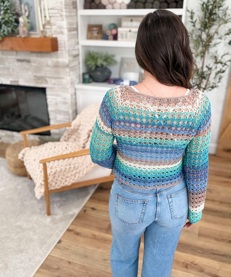 May include: A crocheted top in shades of blue, teal, and brown. The long-sleeved top has a scoop neck and is worn with light blue jeans. The open weave design is visible.