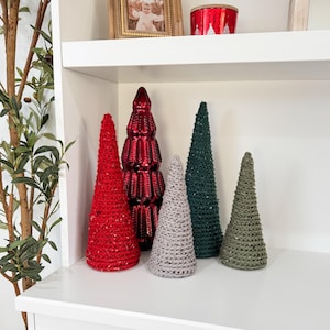 May include: Five decorative Christmas trees on a white shelf. The trees are made of different materials, including red, green, and gray. The trees are all different sizes and shapes.