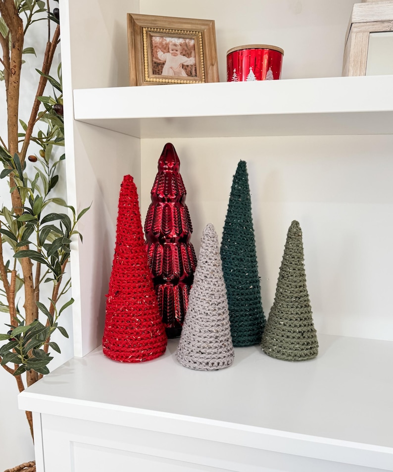 May include: Five decorative Christmas trees on a white shelf. The trees are made of different materials, including red, green, and gray. The trees are all different sizes and shapes.