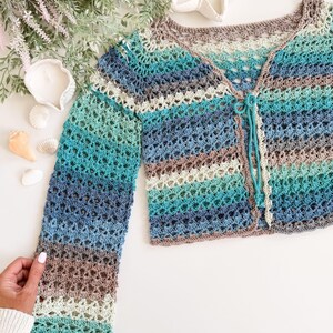 May include: A handmade, open-weave crochet cardigan in shades of blue, teal, and brown. The long-sleeved garment features a button closure and a delicate, airy design. The cardigan is a perfect addition to any wardrobe.