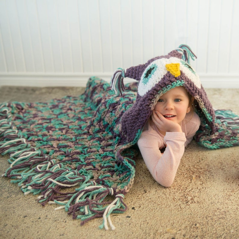 May include: A young girl wearing a purple and green crocheted owl hooded blanket with white and yellow accents. The blanket has fringe around the edges.