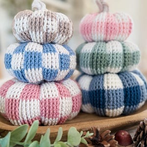 May include: Two stacks of three crocheted pumpkins each, featuring a checkered pattern in various colors including beige, blue, pink, green, and white. Each pumpkin has a small, light-colored stem on top, and they are arranged in two vertical columns.
