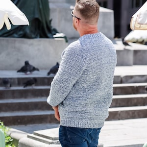 May include: A light blue knit sweater with a crew neck. The sweater has a textured appearance and is worn with blue jeans. The person is standing outdoors near a stone structure.