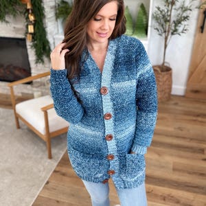 May include: A long, blue crochet cardigan with a V-neck and large brown buttons. The cardigan has two front pockets and a gradient colour scheme, transitioning from light to dark blue. The sleeves are long, and the overall style is casual.
