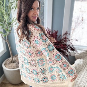 May include: A crocheted cardigan with a granny square pattern in shades of cream, pink, and blue. The cardigan has a long, loose fit and a V-neck.