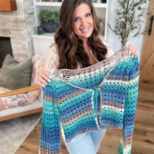 May include: A handmade, open-weave crochet cardigan in shades of blue, teal, brown, and cream. The long-sleeved garment features a button-up front and is held up by a person. The cardigan is a unique, stylish piece.