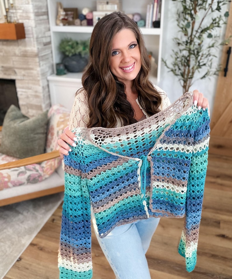 May include: A handmade, open-weave crochet cardigan in shades of blue, teal, brown, and cream. The long-sleeved garment features a button-up front and is held up by a person. The cardigan is a unique, stylish piece.