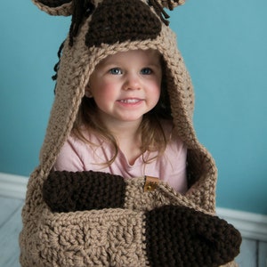 Pu&ograve; includere: Una coperta con cappuccio in crochet marrone e beige con mani in crochet marrone. La coperta presenta un design a testa di cavallo sul cappuccio. Un bambino piccolo indossa la coperta.