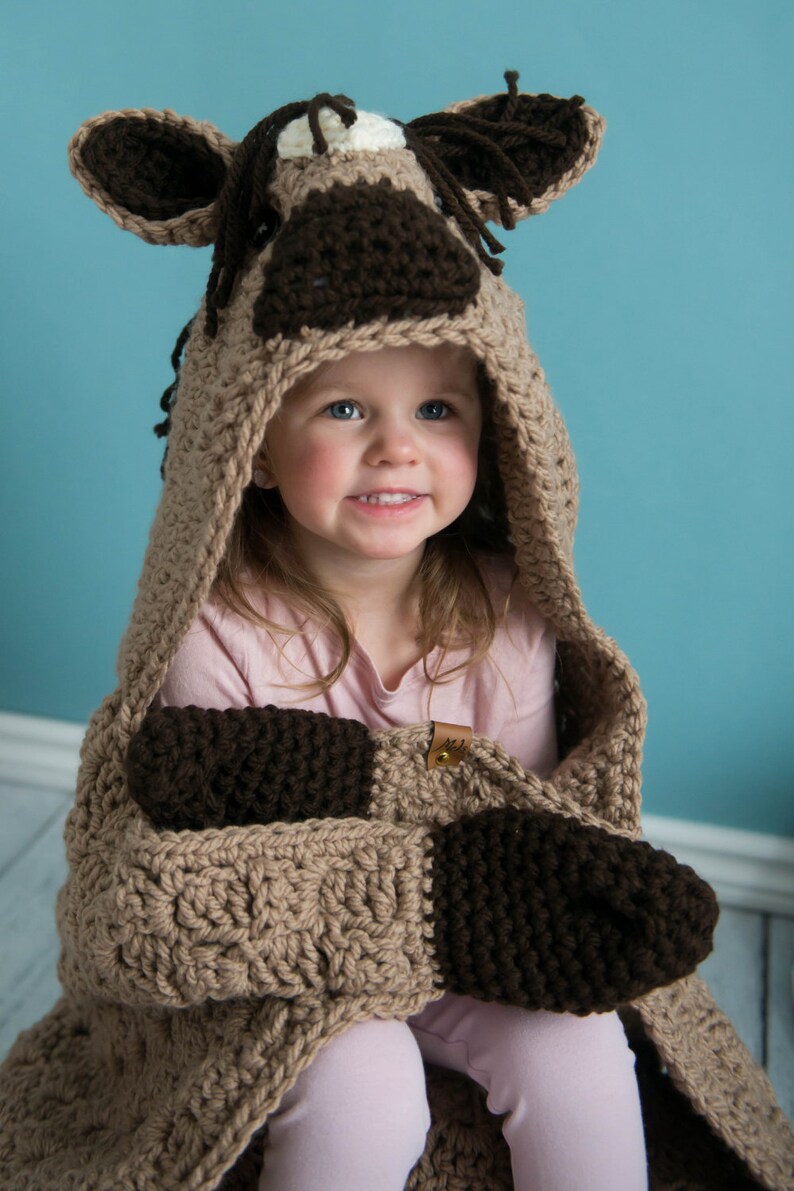 Pu&ograve; includere: Una coperta con cappuccio in crochet marrone e beige con mani in crochet marrone. La coperta presenta un design a testa di cavallo sul cappuccio. Un bambino piccolo indossa la coperta.