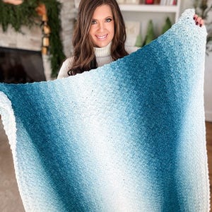 May include: A soft, textured blanket with a gradient of colors, transitioning from white to shades of blue. The blanket appears to be hand-knitted or crocheted, with a dense, plush texture. The overall aesthetic is cozy and inviting, suitable for home decor.