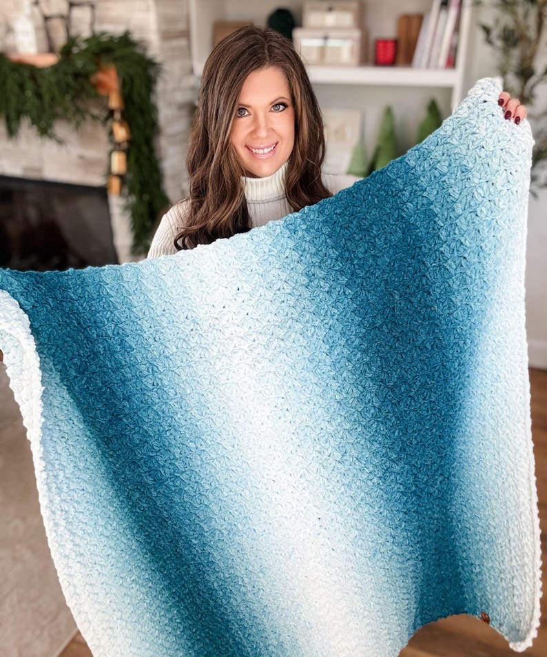 May include: A soft, textured blanket with a gradient of colors, transitioning from white to shades of blue. The blanket appears to be hand-knitted or crocheted, with a dense, plush texture. The overall aesthetic is cozy and inviting, suitable for home decor.
