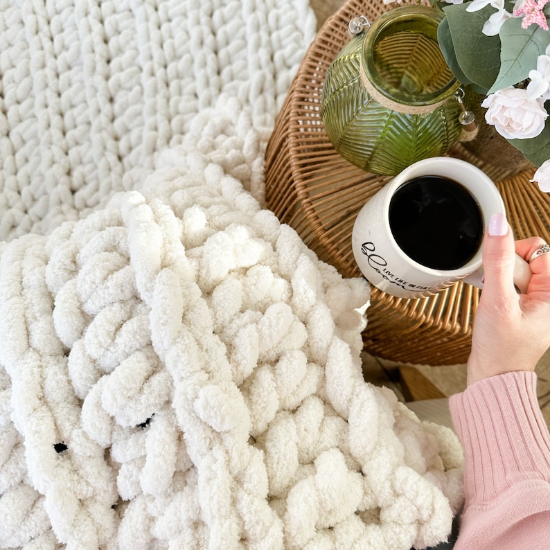 Peut inclure: Une couverture en tricot blanc &eacute;pais avec une tasse de caf&eacute; sur une table en osier &agrave; c&ocirc;t&eacute; d'un vase en verre vert avec des fleurs.