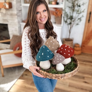 Może przedstawiać: Wiklinowy kosz z pięcioma szydełkowymi dekoracjami w kształcie grzybów. Grzyby mają białe nóżki i kolorowe kapelusze w kolorze morskim, brązowym, czerwonym i beżowym, każdy z białymi kropkami. Kosz spoczywa na zielonym mchu.