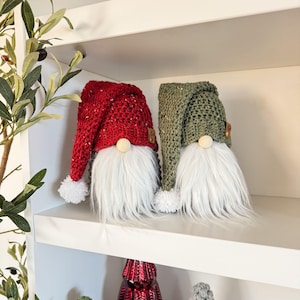 May include: Two crocheted gnomes, one with a red hat and the other with a green hat, sitting on a white shelf. Both gnomes have white fur beards and white pom-poms on their hats.