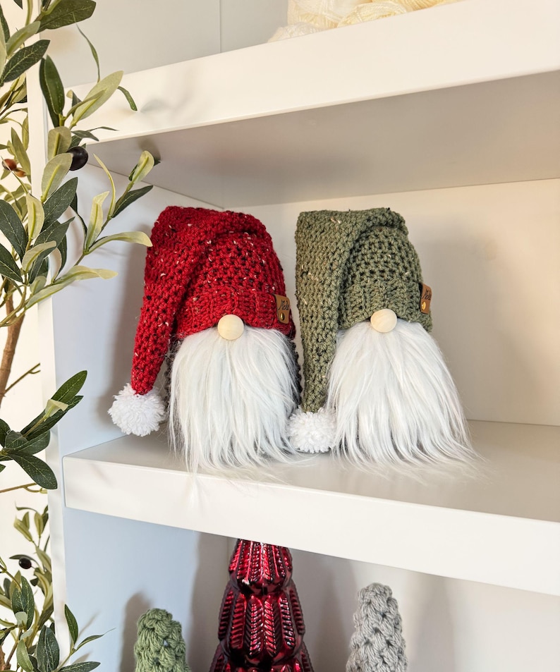May include: Two crocheted gnomes, one with a red hat and the other with a green hat, sitting on a white shelf. Both gnomes have white fur beards and white pom-poms on their hats.