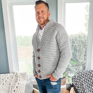 May include: A grey crocheted cardigan with a v-neck and wooden buttons. The cardigan is being worn by a person with a beard and is styled with blue jeans.