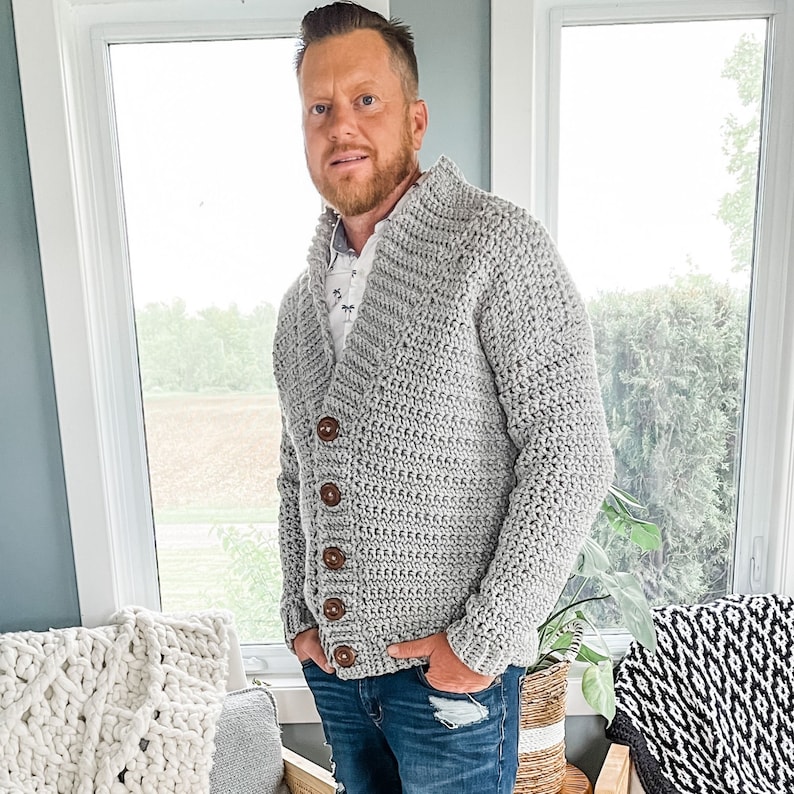 May include: A grey crocheted cardigan with a v-neck and wooden buttons. The cardigan is being worn by a person with a beard and is styled with blue jeans.