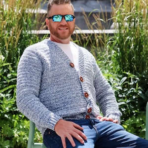 May include: A light blue and white speckled cardigan with brown buttons. The cardigan is worn over a white t-shirt and paired with blue jeans. The person is wearing sunglasses.