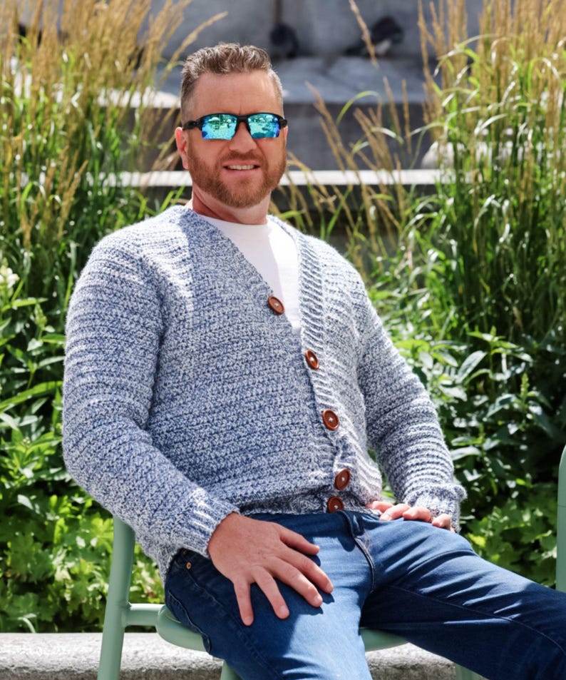 May include: A light blue and white speckled cardigan with brown buttons. The cardigan is worn over a white t-shirt and paired with blue jeans. The person is wearing sunglasses.