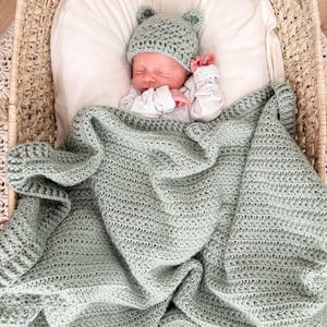 Puede incluir: Una manta de beb&eacute; tejida a ganchillo en color verde salvia con un gorro a juego con orejas de oso. La manta presenta un patr&oacute;n de puntos texturizados y un borde ancho acanalado. El gorro es ajustado con dos peque&ntilde;as orejas redondeadas.