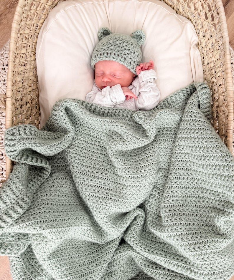 Puede incluir: Una manta de beb&eacute; tejida a ganchillo en color verde salvia con un gorro a juego con orejas de oso. La manta presenta un patr&oacute;n de puntos texturizados y un borde ancho acanalado. El gorro es ajustado con dos peque&ntilde;as orejas redondeadas.