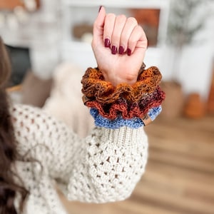 May include: A stack of handmade scrunchies in shades of brown, orange, red, and blue. The scrunchies are made of a textured fabric and are stacked on top of each other. The scrunchies are on a person's wrist.