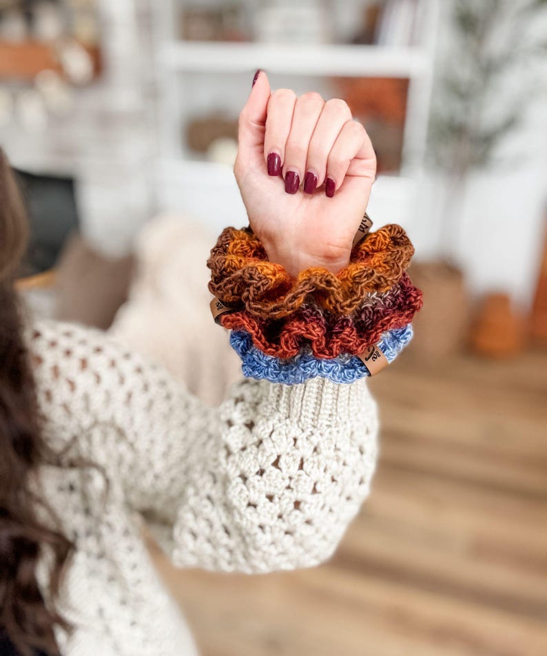 May include: A stack of handmade scrunchies in shades of brown, orange, red, and blue. The scrunchies are made of a textured fabric and are stacked on top of each other. The scrunchies are on a person's wrist.