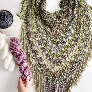 May include: A crocheted shawl in shades of green, brown and grey. The shawl has a fringed border and is made with a textured stitch. Three balls of yarn are shown in the image, one white, one pink and one grey.