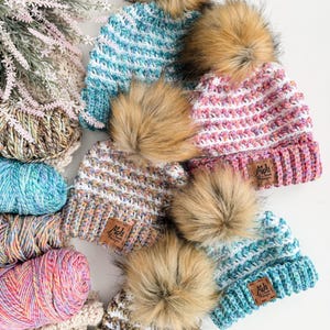 Può includere: Una collezione di cappelli lavorati a maglia colorati, fatti a mano, con pompon in finta pelliccia. I cappelli presentano un bordo a coste e un motivo a punto strutturato in vari colori, tra cui blu, rosa e marrone. Ogni cappello ha una piccola etichetta rettangolare con il testo "Mds".