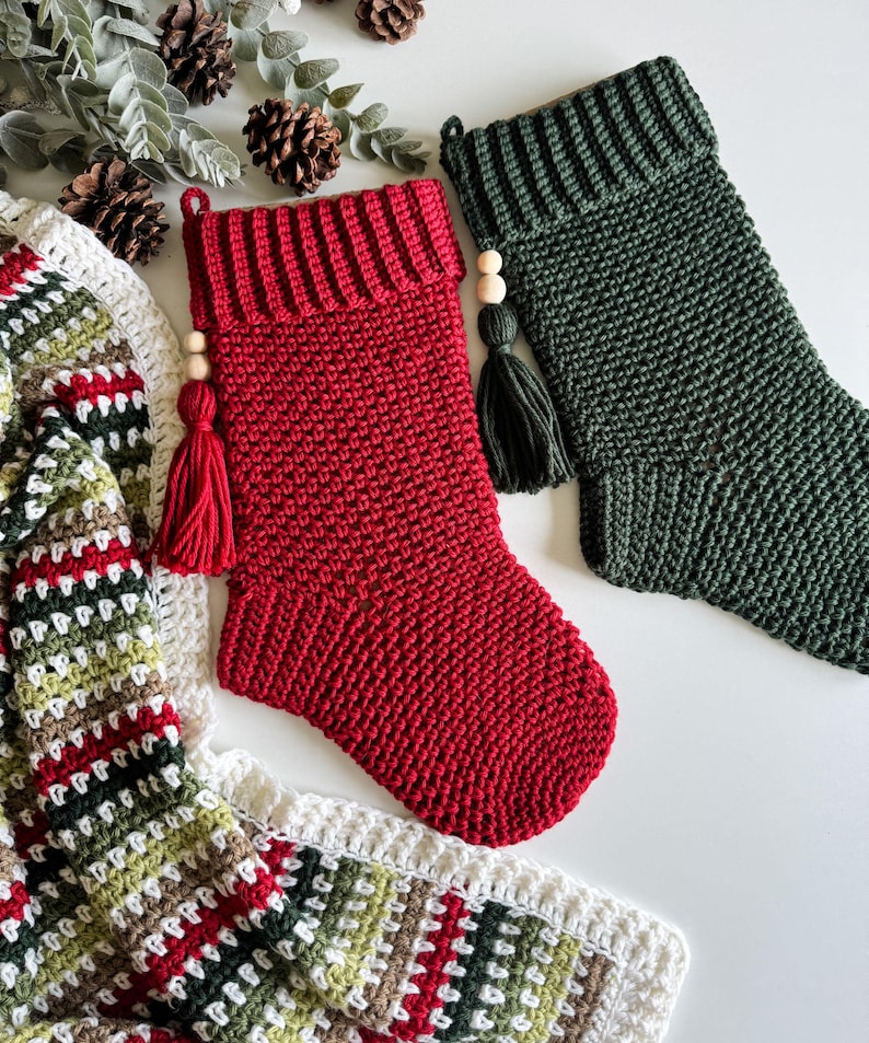 May include: Two handmade crochet Christmas stockings, one red and one green, with tassels and wooden beads. A crocheted blanket with red, green, and white stripes is also visible. The stockings are a festive holiday decoration.