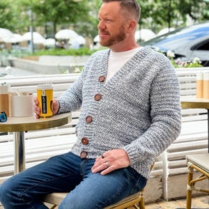 May include: A light blue and white knit cardigan with large brown buttons. The cardigan is worn over a white t-shirt and paired with blue jeans. The person is holding a yellow coffee cup with black text.