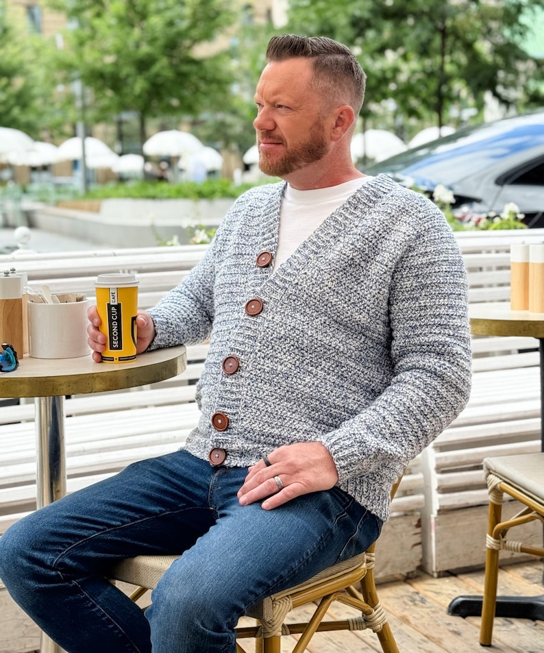 May include: A light blue and white knit cardigan with large brown buttons. The cardigan is worn over a white t-shirt and paired with blue jeans. The person is holding a yellow coffee cup with black text.