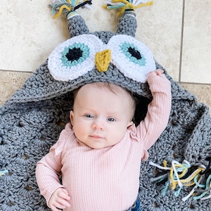 May include: A baby wearing a gray crocheted owl hooded blanket with white and teal eyes. The blanket has tassels in yellow, teal, and white.