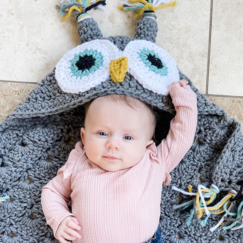 May include: A baby wearing a gray crocheted owl hooded blanket with white and teal eyes. The blanket has tassels in yellow, teal, and white.