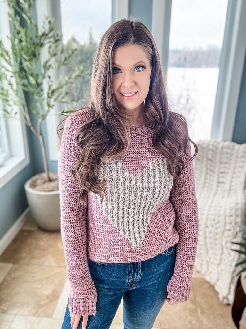 Puede incluir: Un su&eacute;ter de crochet malva con un dise&ntilde;o de coraz&oacute;n blanco. El su&eacute;ter tiene mangas largas y un ajuste holgado.