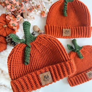 May include: Three orange knit beanies shaped like pumpkins, each featuring a green crocheted stem and leaf on top. Each hat has a ribbed cuff and a square tan leather-like patch with the text MJs off the hook designs stitched on the front.